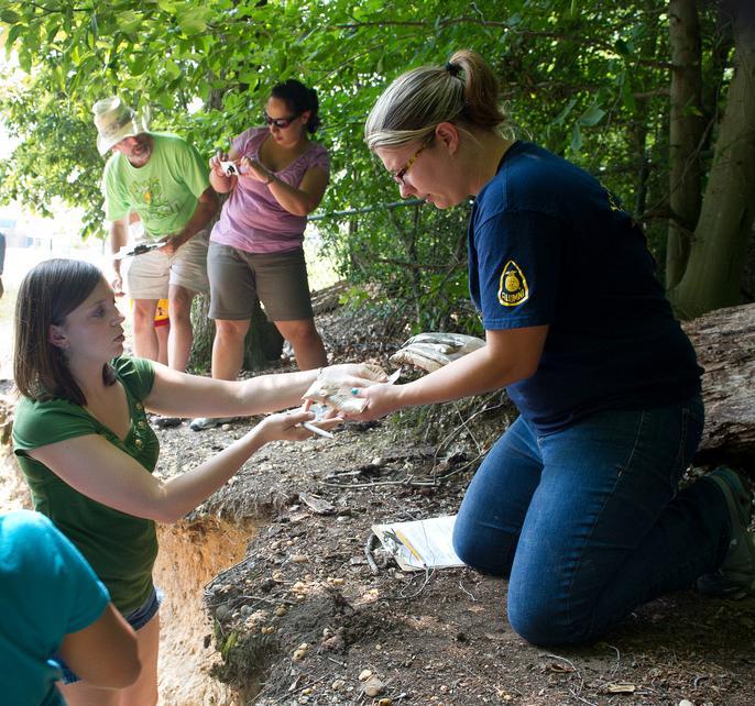 Woman kneeling on dirt giving student a sample, two people in background taking photos