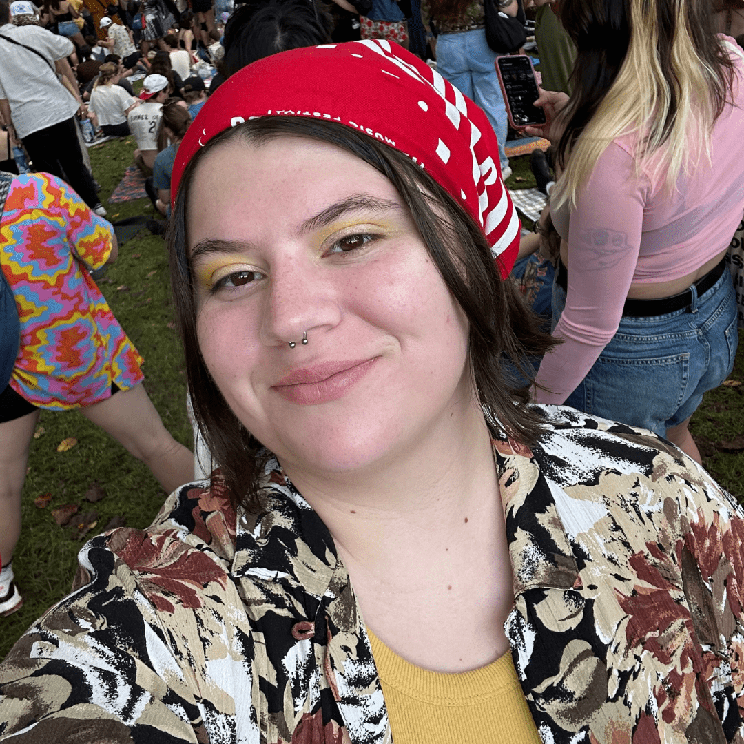 Sophie is standing in a crowd and smiling at the camera, wearing a red bandana, yellow top, and yellow eyeshadow.