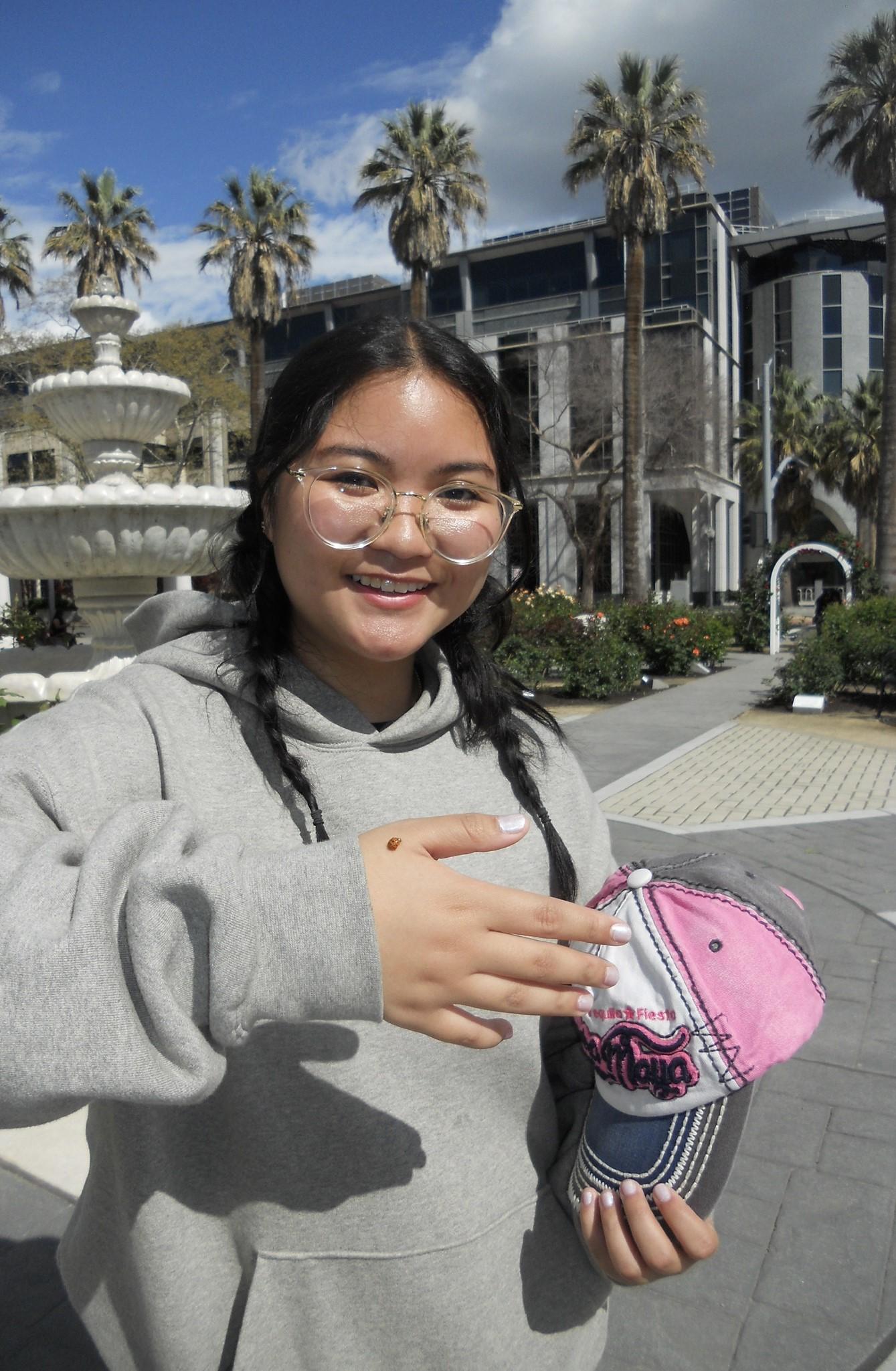 Happy student holding pink baseball cap in front of palm trees and fountain. Showcasing ladybug that landed on her wrist.