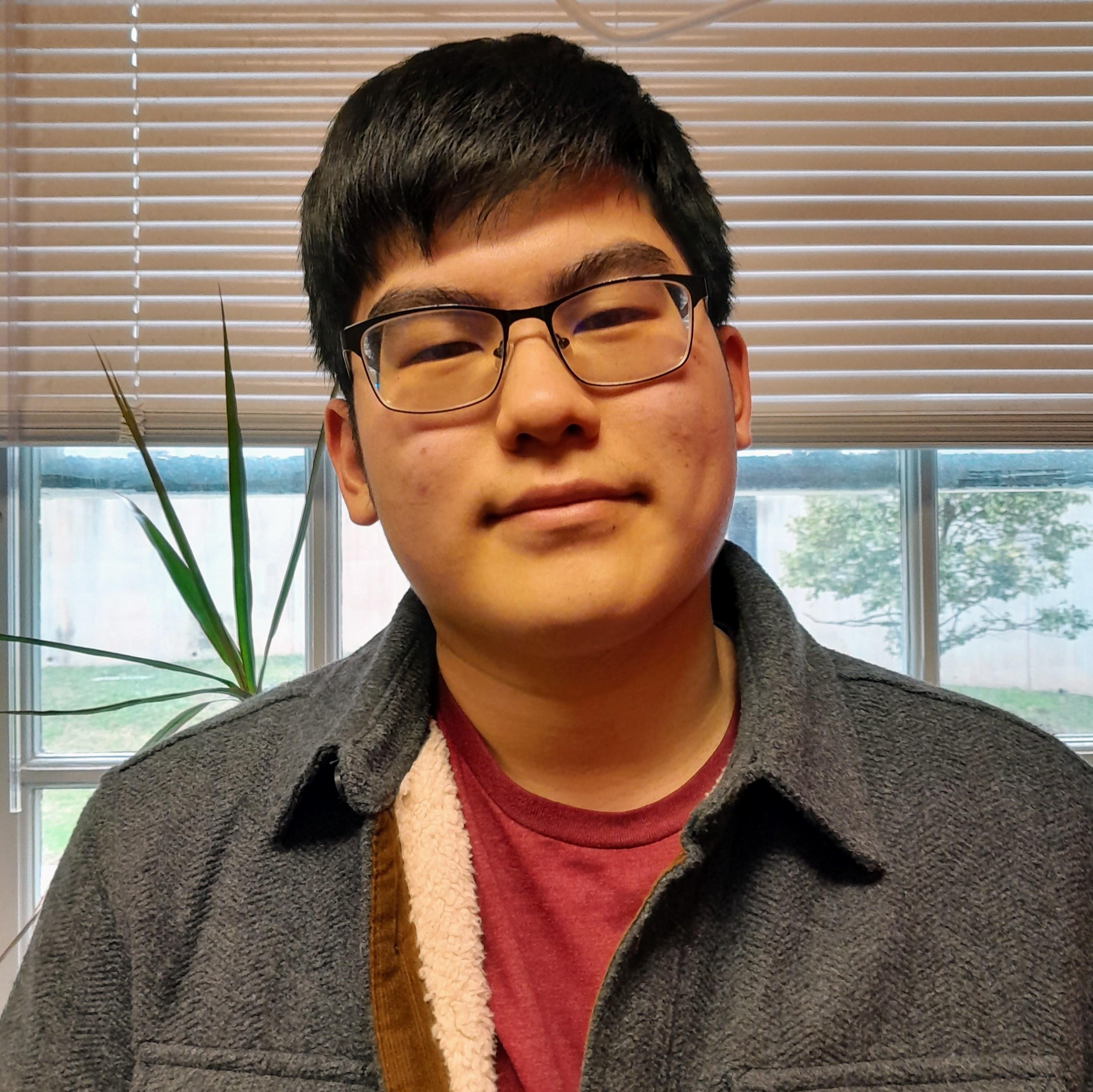 Jason is smiling and standing in front of a window, wearing a grey jacket over a red tee.