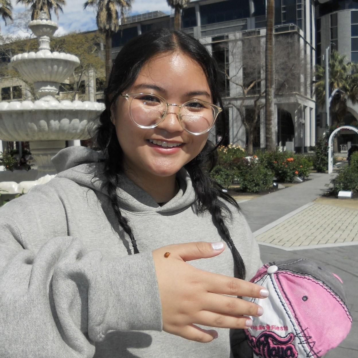 Grazelle is standing in front of a fountain and palm trees, smiling with a lady bug on her hand